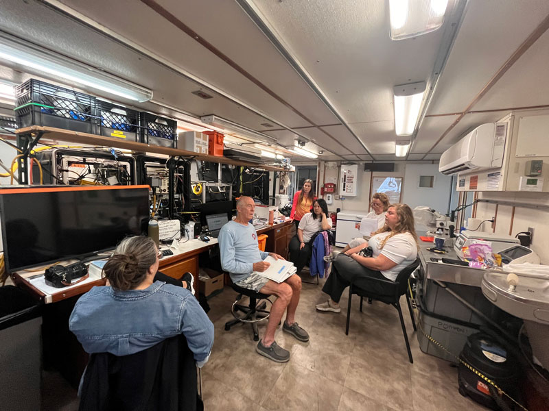 John Reed, co-chief scientist, addresses the science crew prior to a dive during Exploration of Deepwater Habitats off Puerto Rico and the U.S. Virgin Islands for Biotechnology Potential. Clockwise from left to right: Megan Conkling, John Reed, Carol Kim, Courtney Brooks, Cristina Diaz and Kirstie Francis.