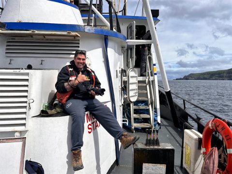 The image depicts a smiling man sitting on the edge of a section of a boat. He is giving a thumbs-up gesture while holding a camera. The man is wearing a black jacket, a red life vest, gray sweatpants, and brown work boots. The weather appears partly cloudy. The boat has a predominantly white structure with blue trim. Behind the man, there is a set of white stairs leading to another level of the boat. The open water is visible on the right side of the image, with a coastline featuring green hills in the distance. A red life preserver ring is mounted on the railing at the edge of the boat.